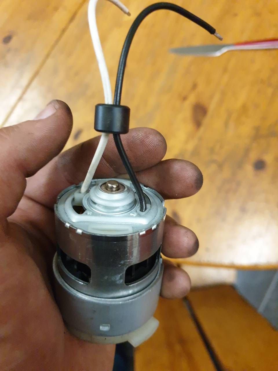 Eine Hand hält einen kleinen grauen zylindrischen Motor mit angeschlossenen Kabeln über einem Holzboden.