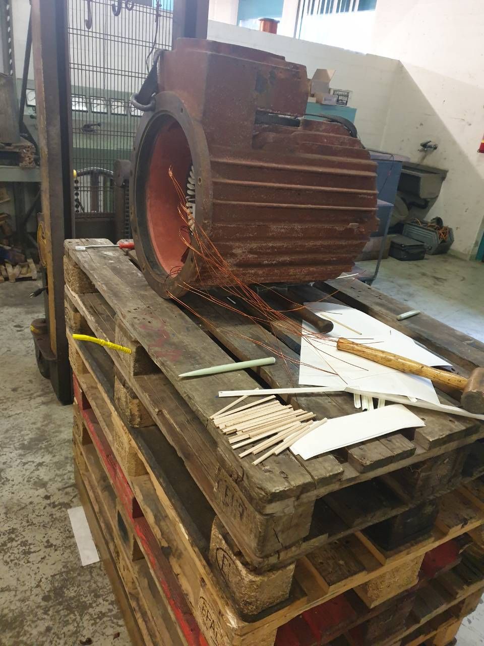 Ein grosser, rostiger Industriemotor oder Generator steht auf einer Holzpalette in einer Werkstatt, daneben Werkzeuge und Schrott.
