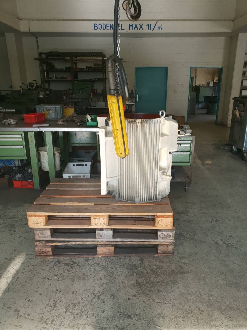 Ein gelber Hebegurt hängt eine weisse Maschine über Holzpaletten in einer Industriewerkstatt.