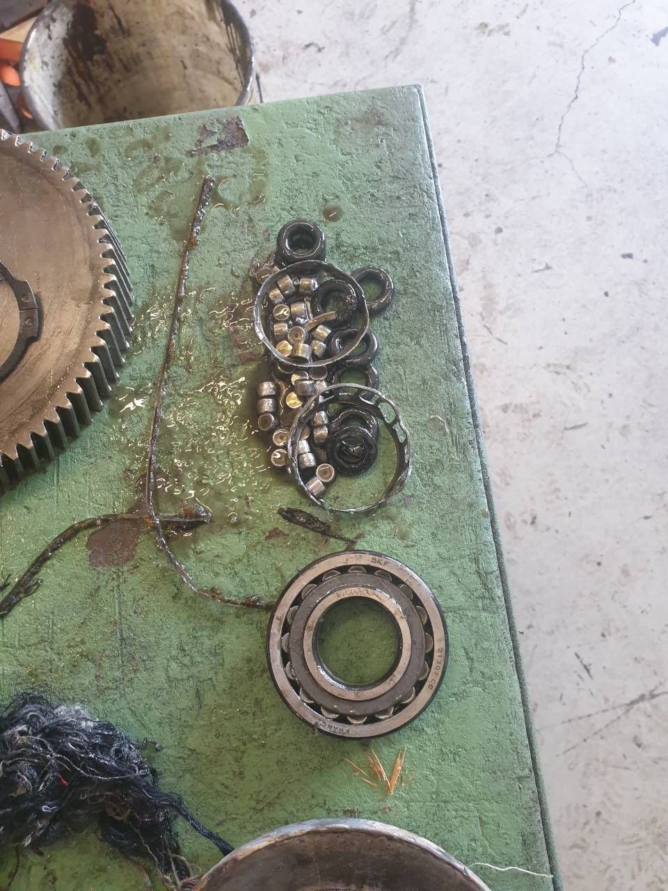 Grüne Werkbank mit verstreuten Metalllagern, Zahnrädern und öligen Maschinenteilen.