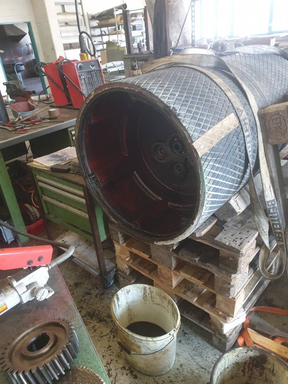 Grosse Metalltrommel auf Holzpaletten in einer vollgestellten Werkstatt, daneben Werkzeuge und Eimer.