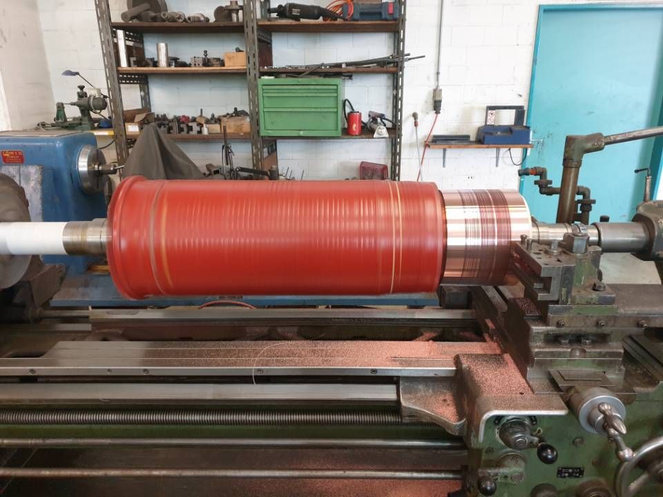 Ein rotes, zylindrisches Werkstück ist in einer Industriewerkstatt auf einer Drehbank montiert; im Hintergrund sind Regale und Werkzeuge zu sehen.