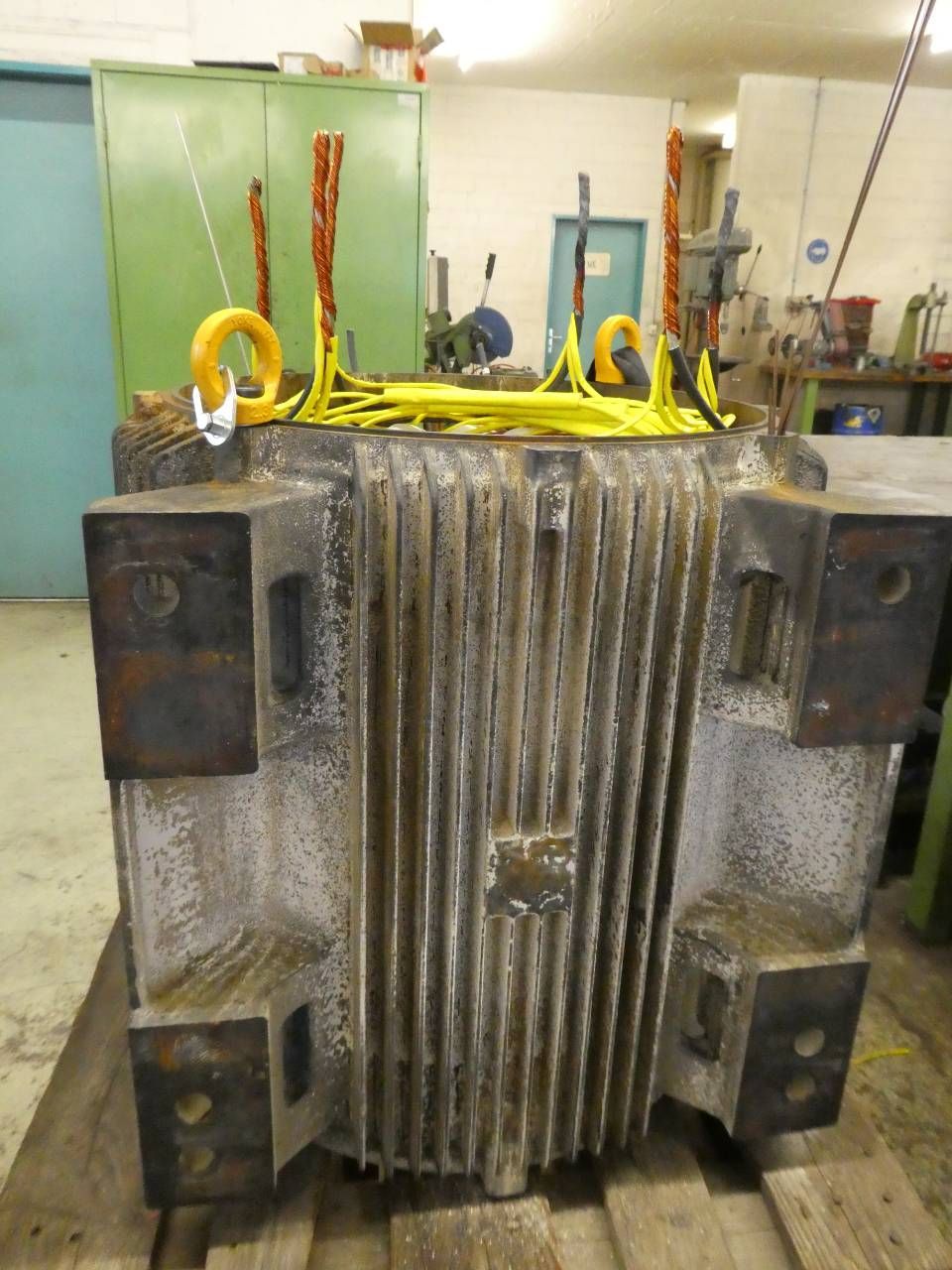 Industrieller Metalltransformator mit Kühlrippen und gelben Hebebändern in einer Werkstatt.