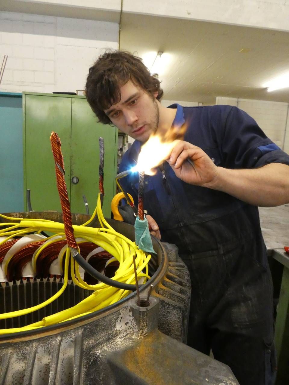 Eine Person in einer Werkstatt bearbeitet mit einer Flamme ein grosses Kabelbündel mit gelben Drähten.