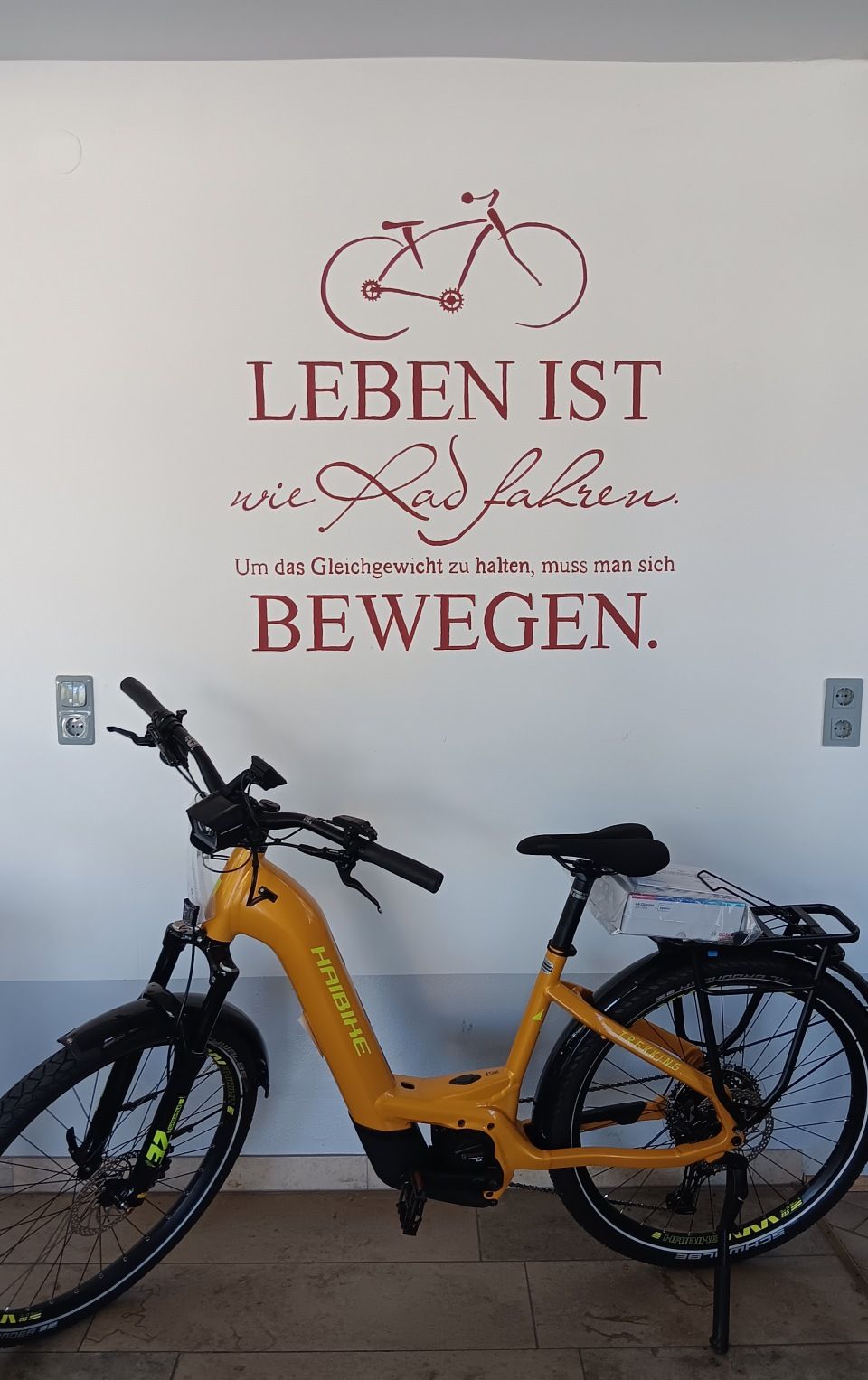 Ein gelbes Elektrofahrrad lehnt an einer Mauer mit einem deutschen Schild „Leben ist bewegen“.