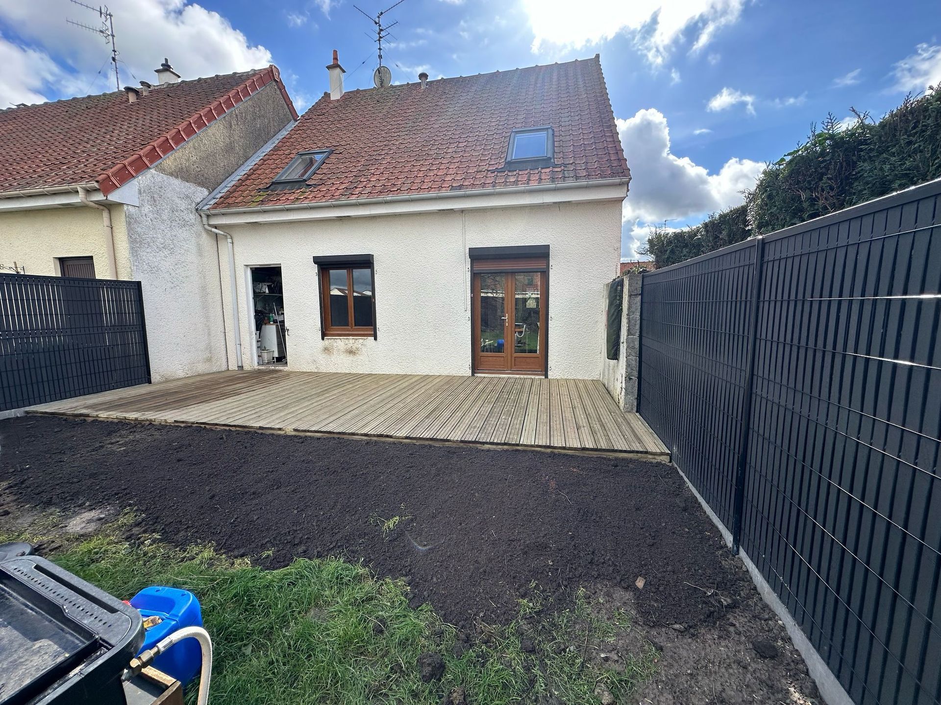 Vue arrière d'une petite maison avec une terrasse en bois, des clôtures noires et un ciel bleu nuageux.
