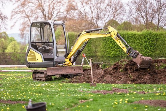 Une petite pelleteuse creuse la terre dans un jardin gazonné, avec une haie et des arbres en arrière-plan.