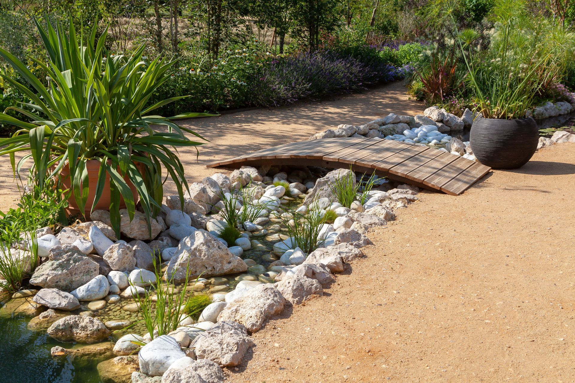 Bassin de jardin avec rochers blancs, petit pont et plantes en pot au bord d'un chemin sablonneux.