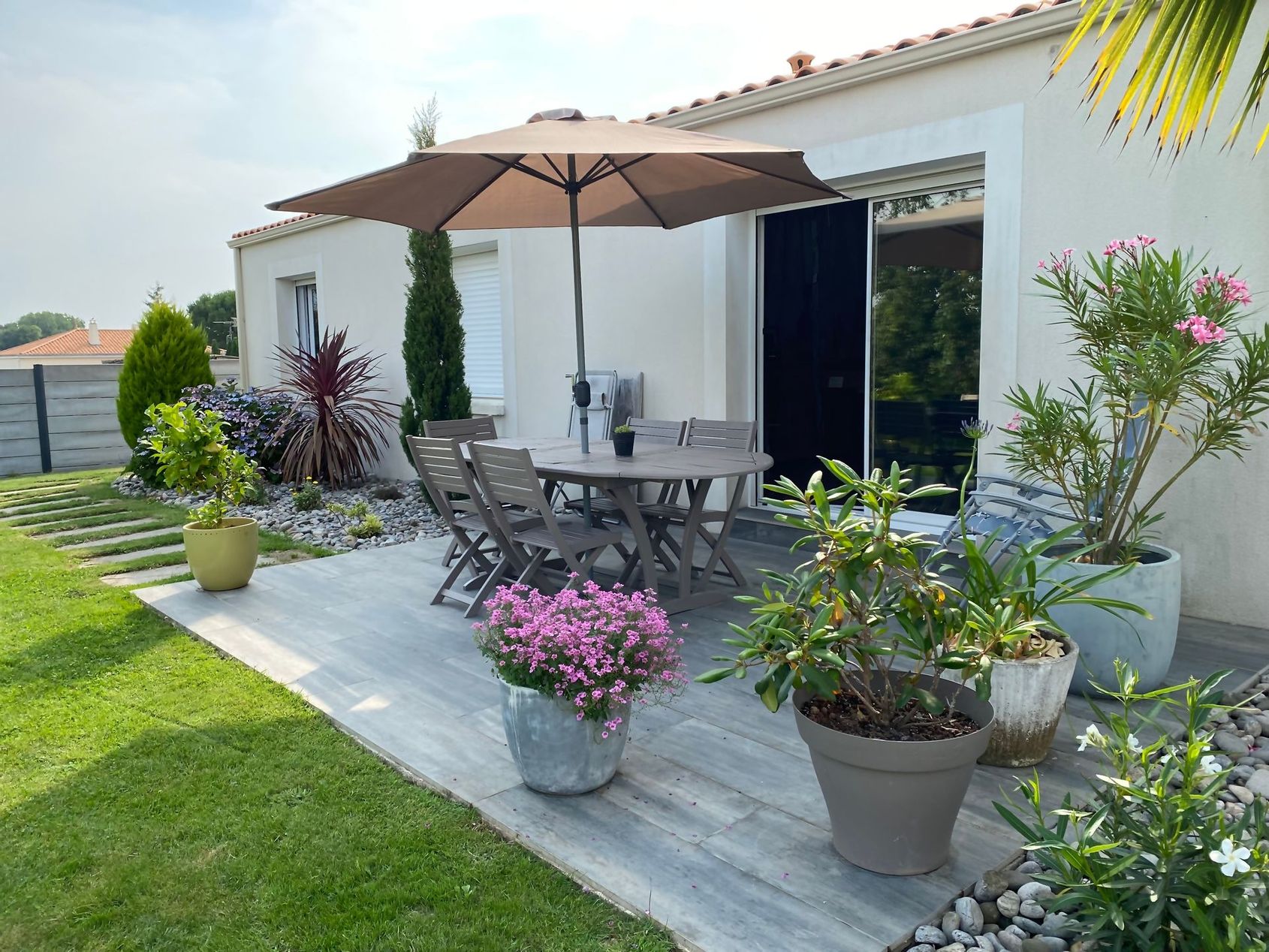 Terrasse avec table, chaises, parasol et fleurs en pot à côté d'une maison blanche et d'une pelouse verte.