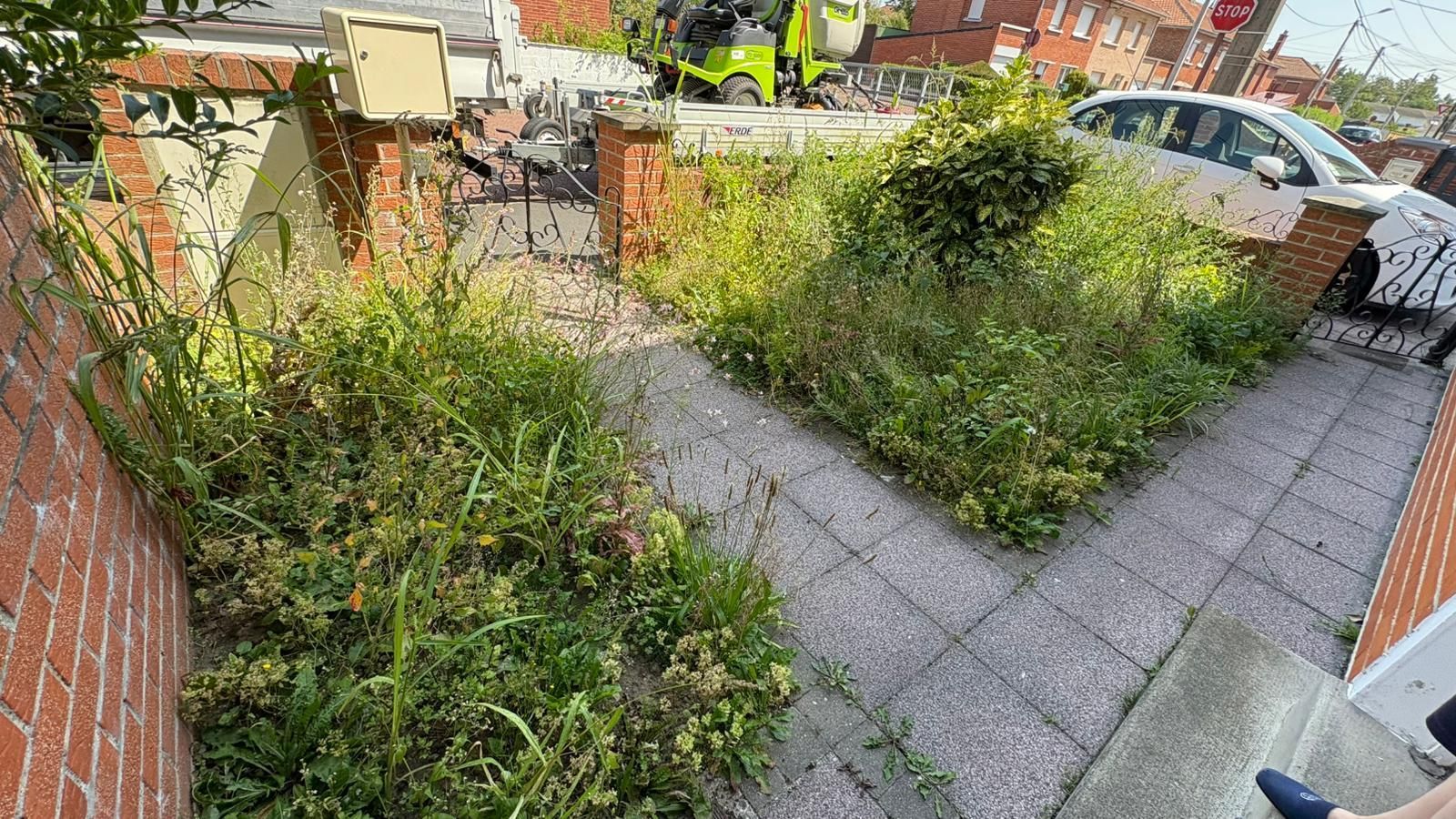 Jardin de devant envahi par la végétation, avec murs de briques, pavés et voitures garées derrière le portail.