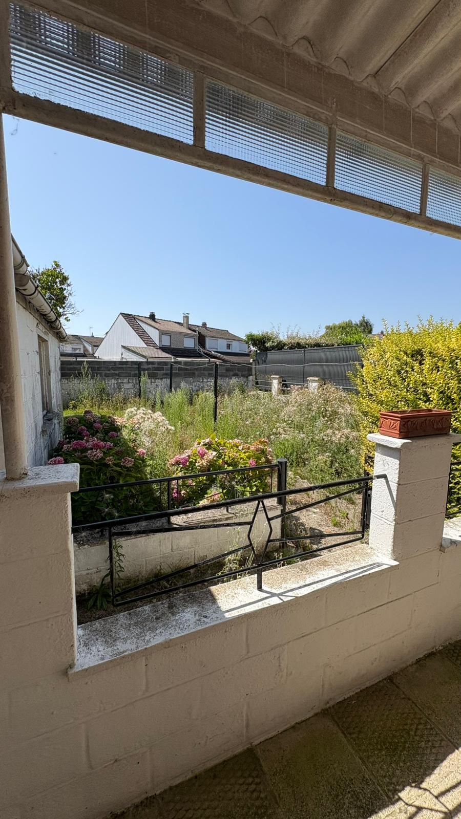 Balcon ombragé donnant sur un jardin lumineux et les maisons voisines.