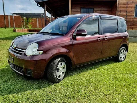 Un pequeño coche rojo está aparcado en el césped delante de una casa de madera.