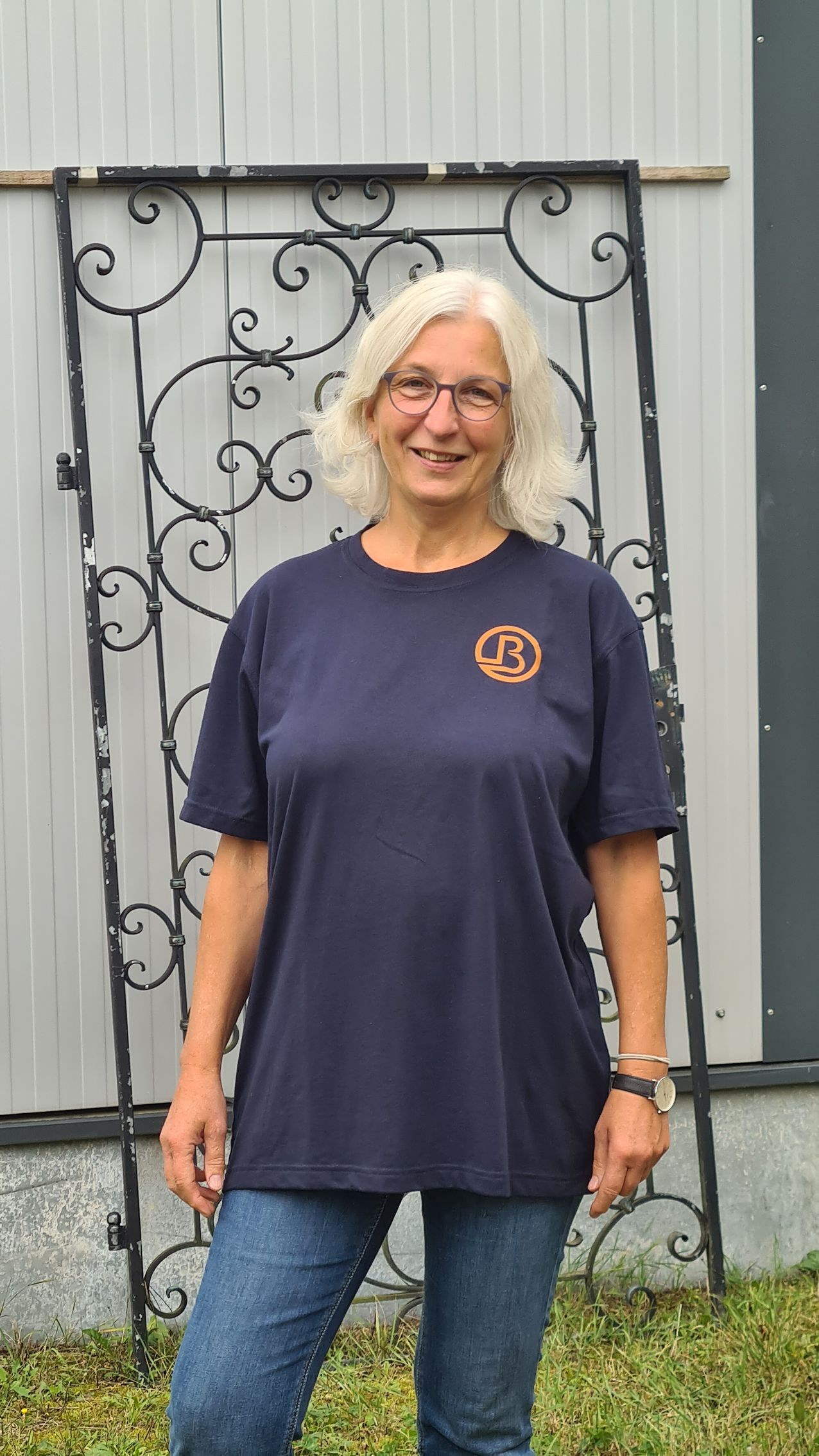 Eine Frau steht im Freien vor einem kunstvollen Eisengitter, trägt ein dunkelblaues T-Shirt und Jeans 