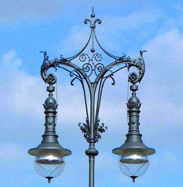 Eine kunstvoll verzierte schwarze Straßenlaterne mit zwei hängenden Laternen vor blauem Himmel