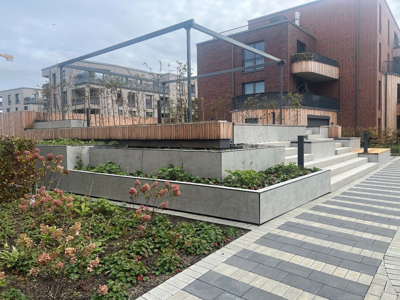 Modernes Backsteingebäude mit gestuften Pflanzbeeten, Metallpergola und gestreiftem Gehweg auf einem begrünten Platz