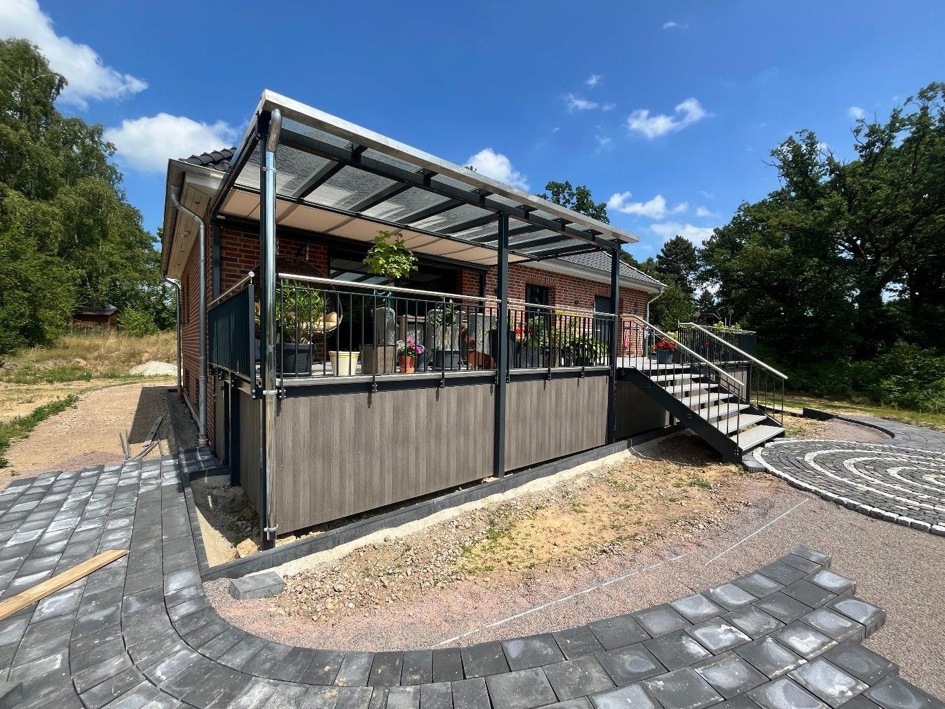 Überdachte Terrasse im Freien mit Metallgeländer, Treppe und Kopfsteinpflasterwegen in einer parkähnlichen Landschaft
