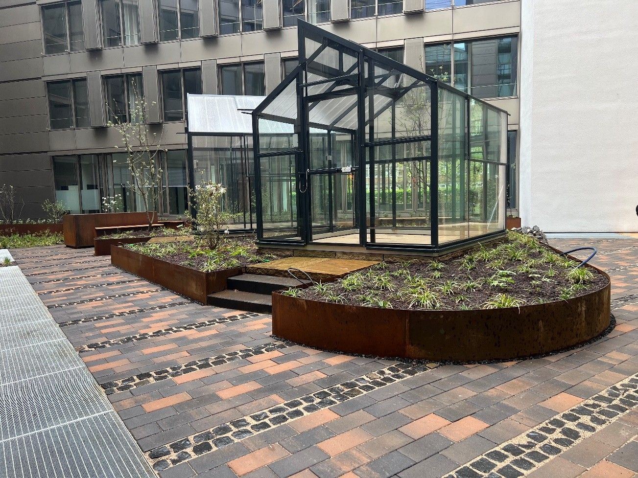 Urbaner Innenhof mit Hochbeeten und einem Gewächshaus aus Glas auf einer gemusterten Ziegelterrasse