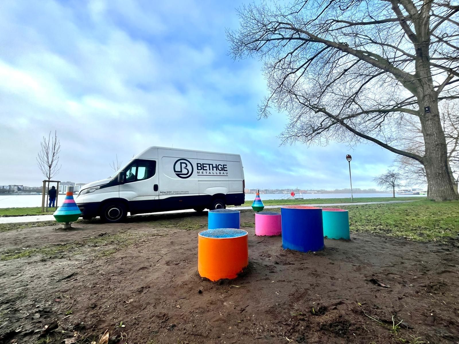 Weißer Lieferwagen neben bunten Fässern an einem Flussufer unter bewölktem Himmel