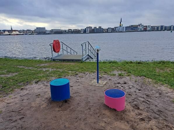 Sandstrand mit zwei blauen und rosa Eimern, einem Steg am Seeufer und Stadtgebäuden auf der anderen Wasserseite.
