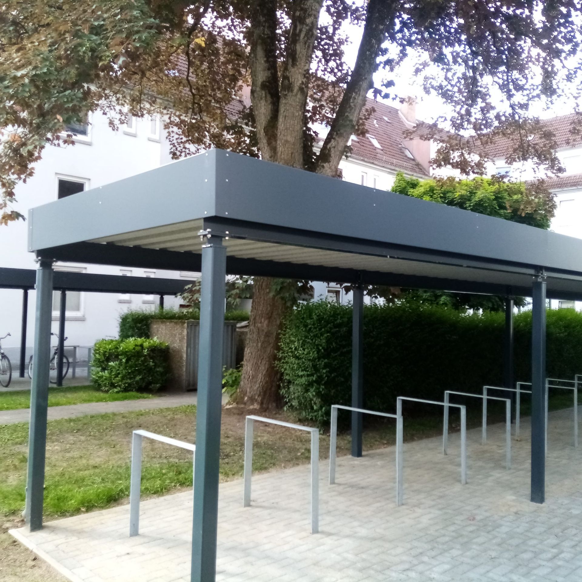 Überdachter Fahrradparkplatz mit Metallständern und dunkelgrauem Dach neben einem Gebäude und Bäumen