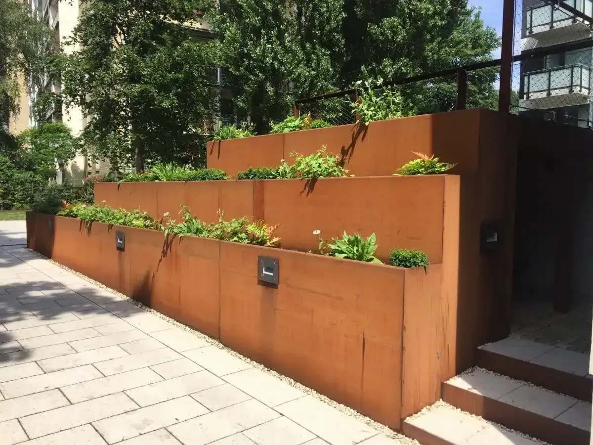Rostfarbene, terrassenförmig angelegte Pflanzmauer mit Grünpflanzen neben einem Gehweg und Treppen in einem Innenhof