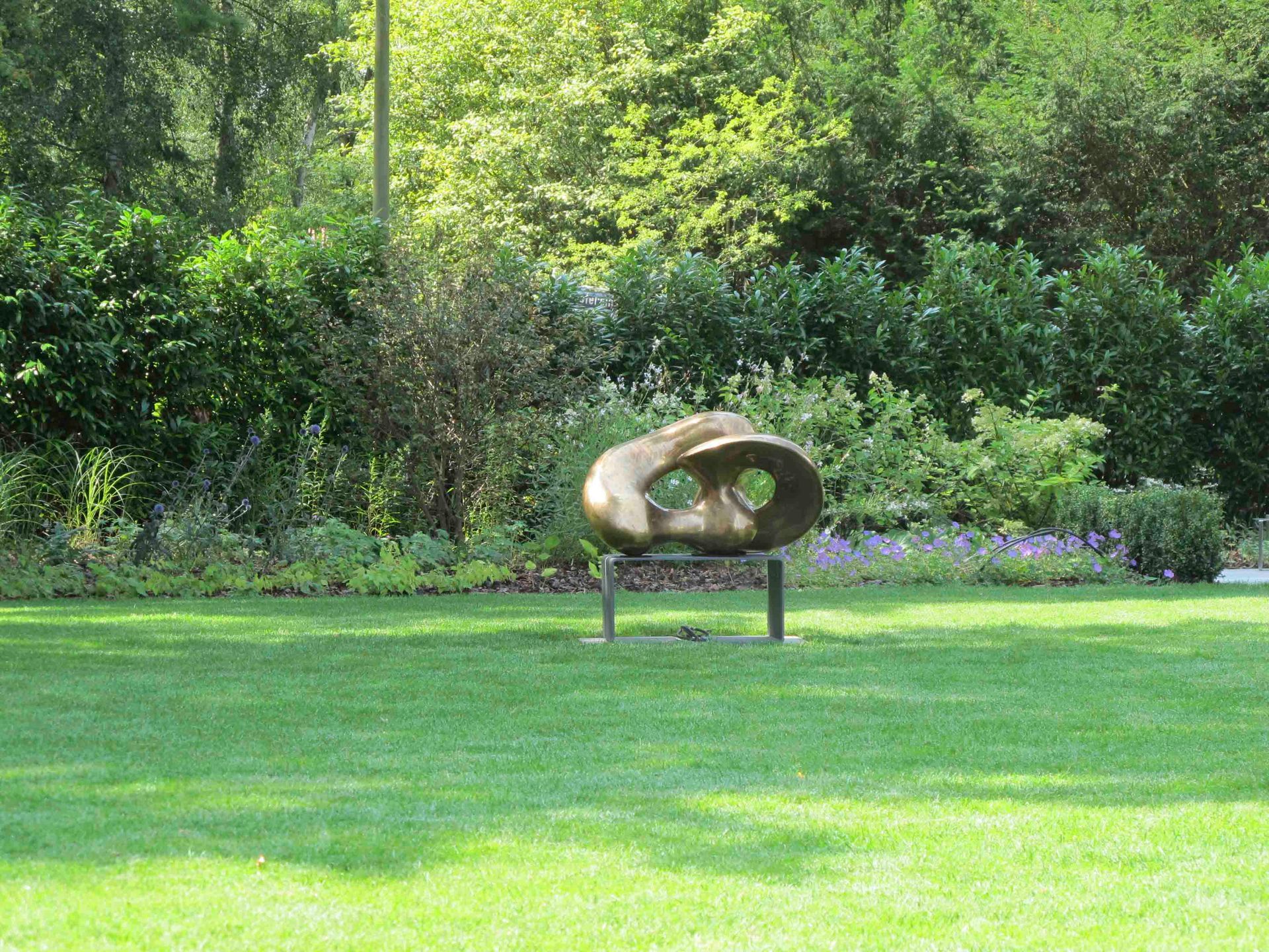 Skulptur auf einem Sockel in einem grasbewachsenen Park, im Hintergrund Bäume und Sträucher.