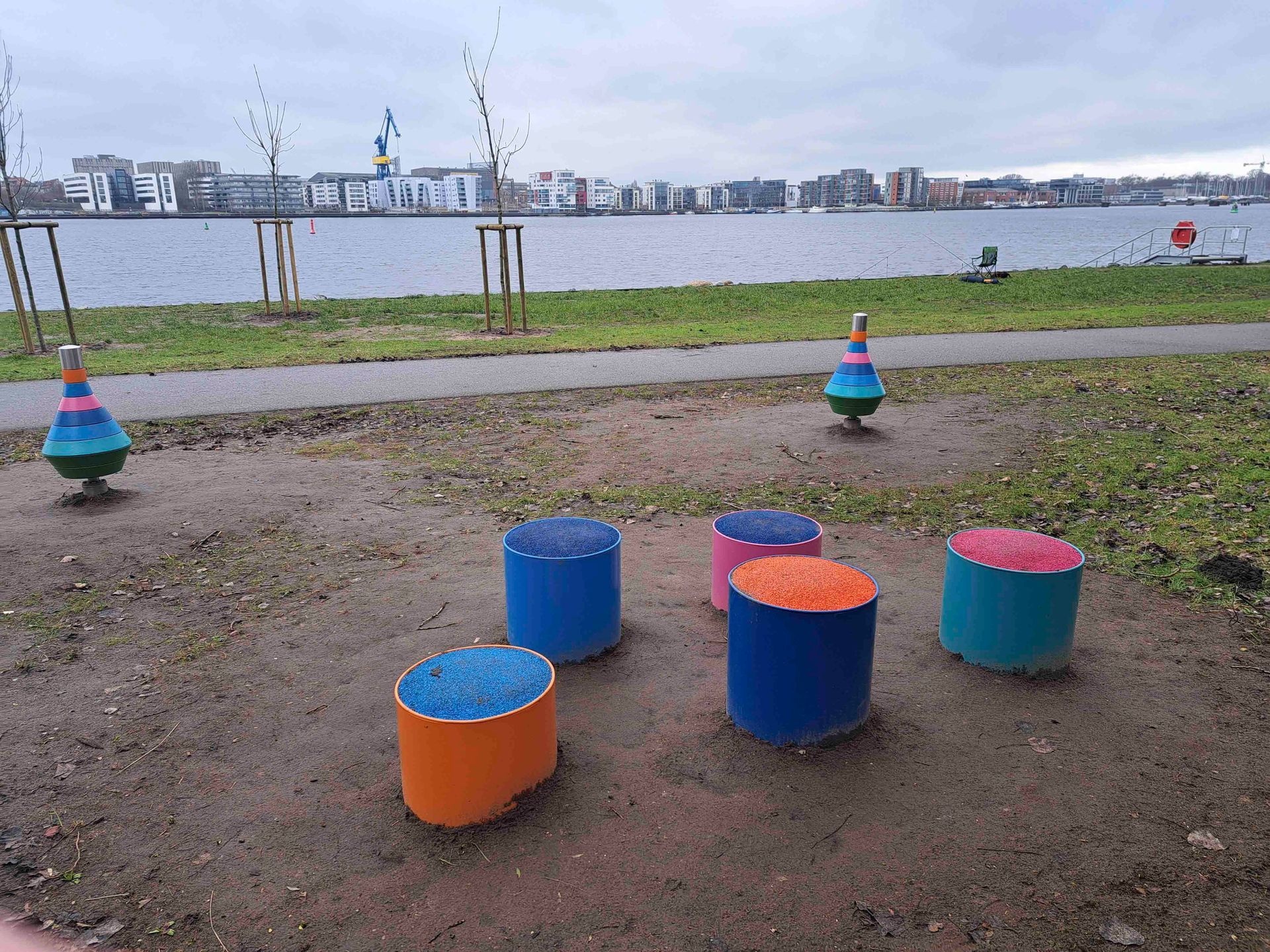 Bunte blaue und orangefarbene Hocker auf einem Kiesweg am Wasser mit der Skyline der Stadt in der Ferne