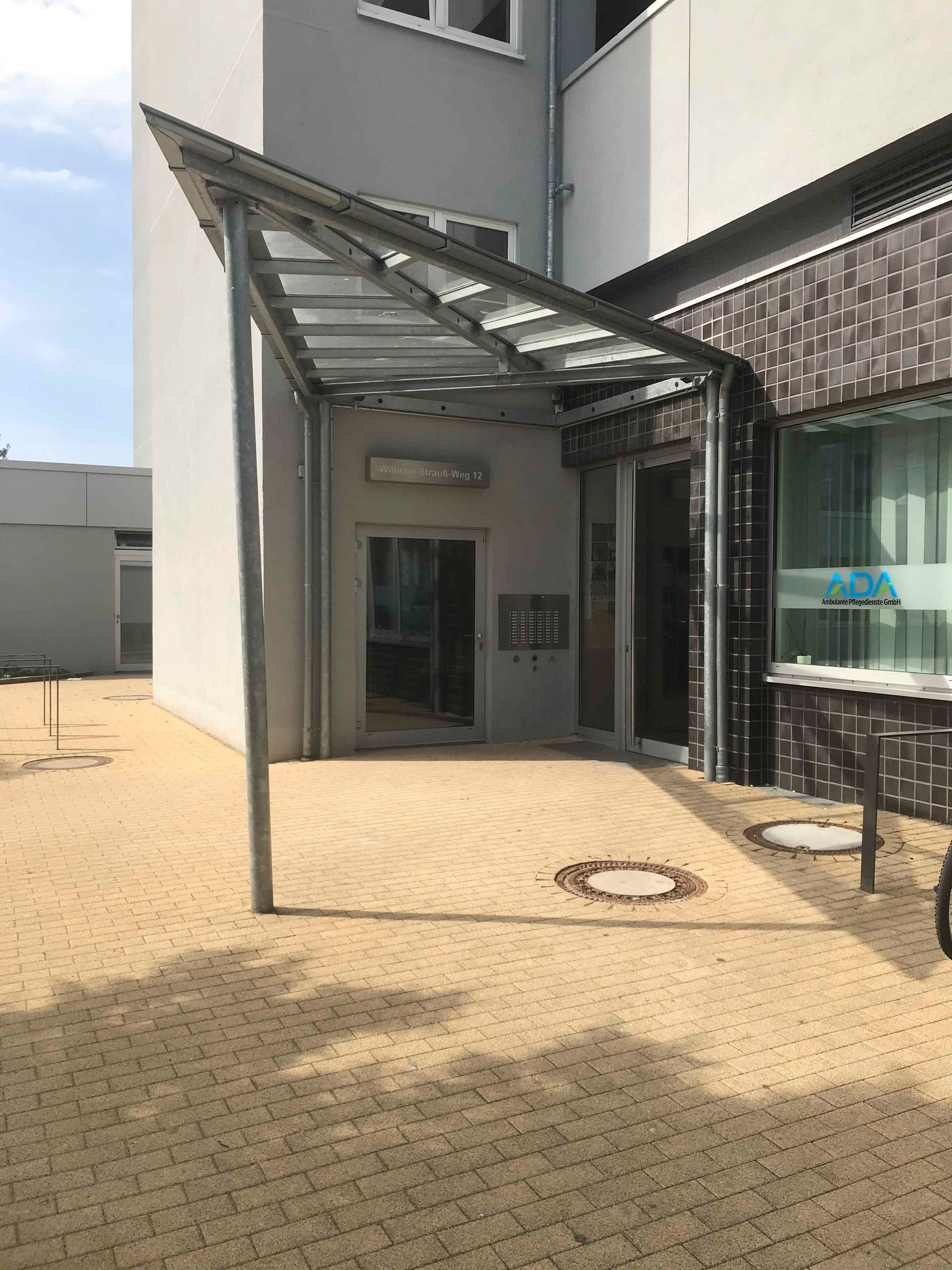 Überdachter Eingang mit Glasvordach neben einem Gebäude auf einem gepflasterten Innenhof