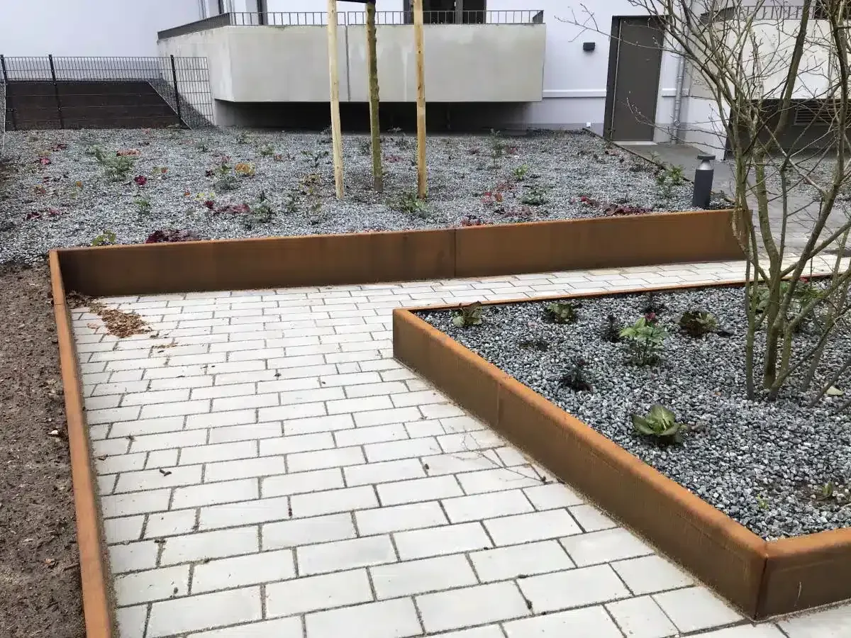 Gartenweg mit hellen Ziegelsteinen und erhöhten Holzkästen, die mit Kies und spärlichen Sträuchern gefüllt sind.