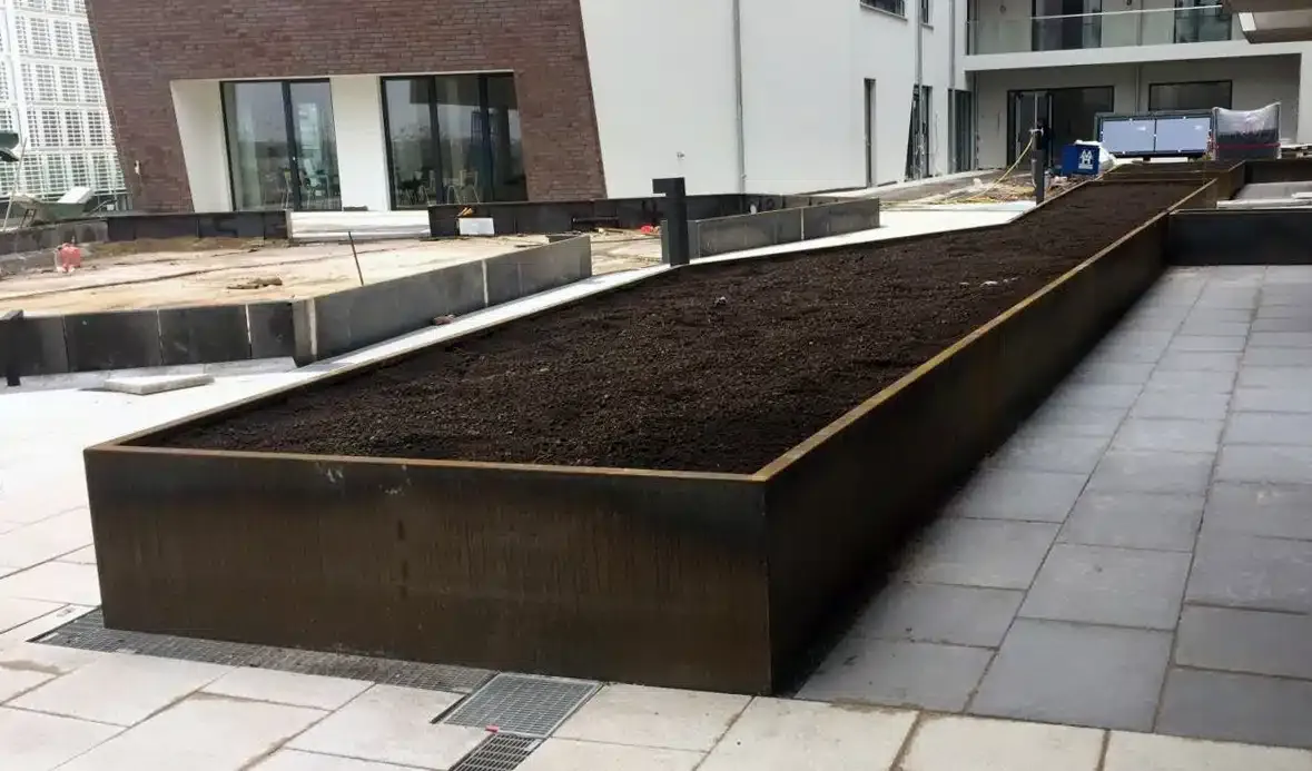 Ein länglicher, rechteckiger Hochbeetkasten, gefüllt mit Erde, steht auf einem gepflasterten Stadtplatz.