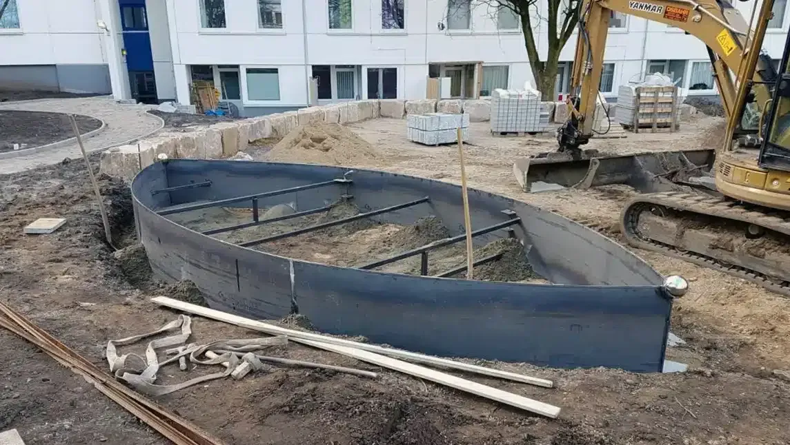 Baustelle mit Bagger und gebogener schwarzer Stützmauer vor einem Gebäude