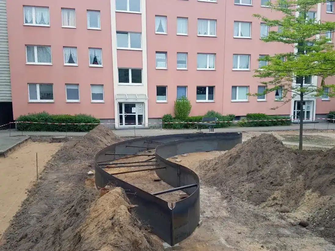 Aushubbereich mit Erdhaufen und schwarzer Einfassung vor einem rosafarbenen Wohnhaus