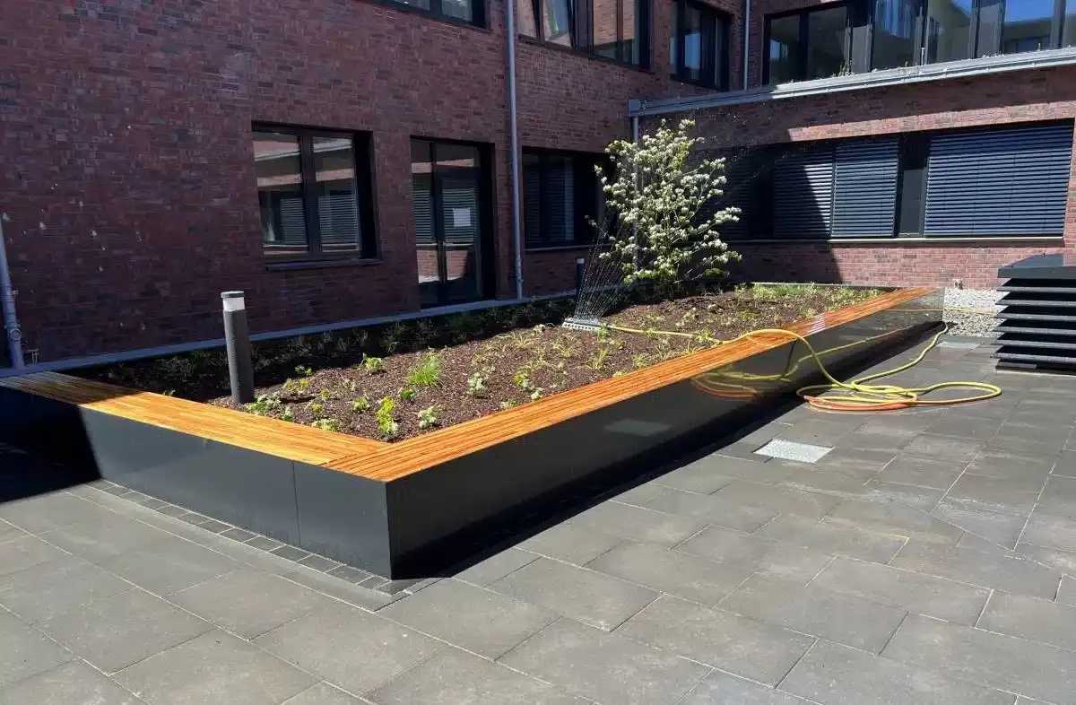 Hochbeet mit Blumen neben einem Backsteingebäude und einem gepflasterten Gehweg