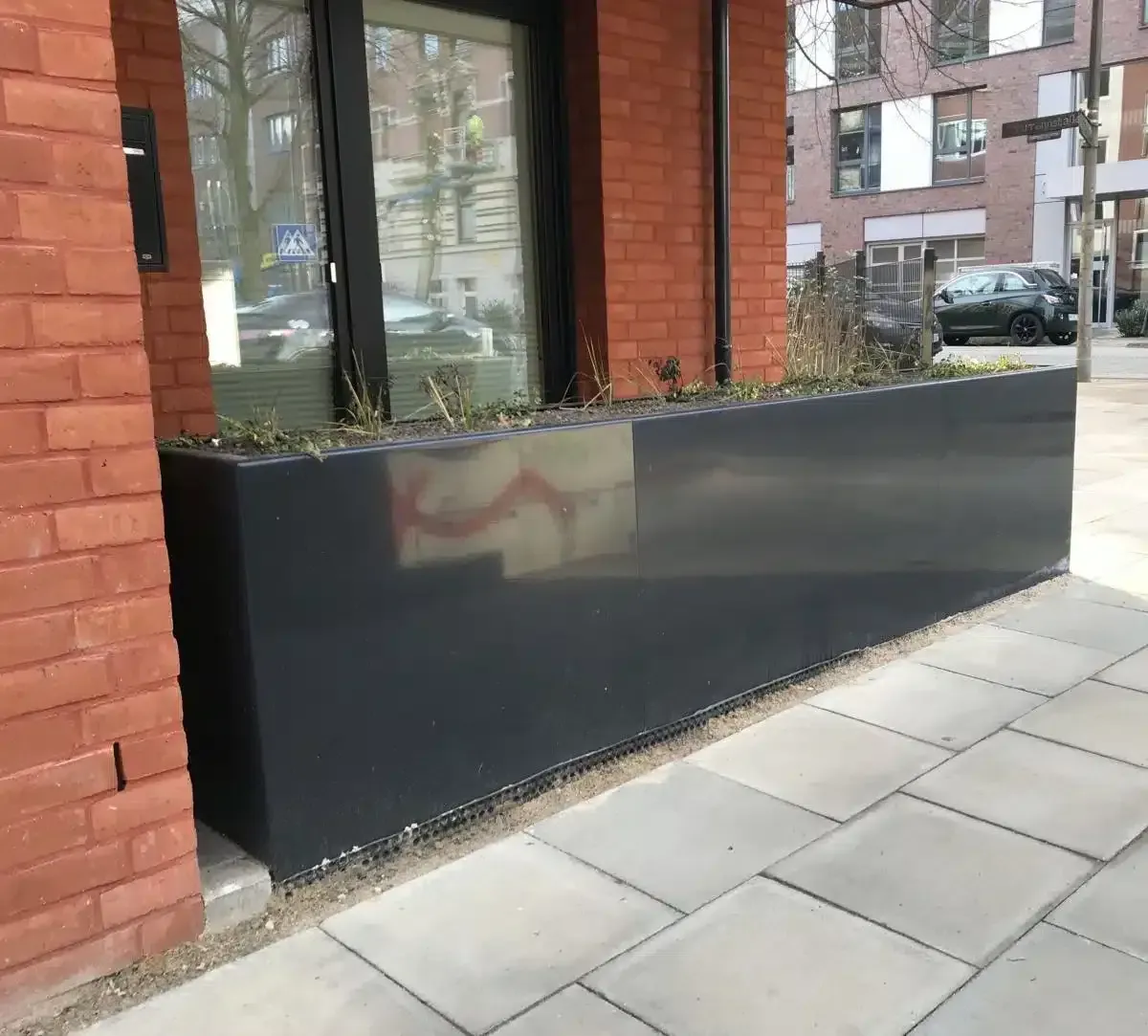 Ein schwarzer Pflanzkasten neben einem Backsteingebäude und einem Gehweg, im Hintergrund Fenster und geparkte Autos.