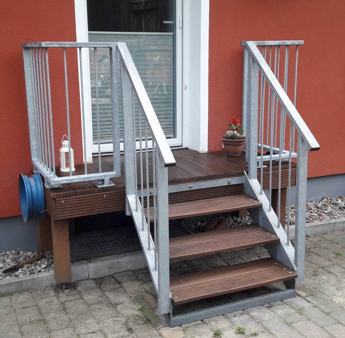 Kleine Holzveranda mit Metallgeländer und Treppe, die zu einem roten Hauseingang führt