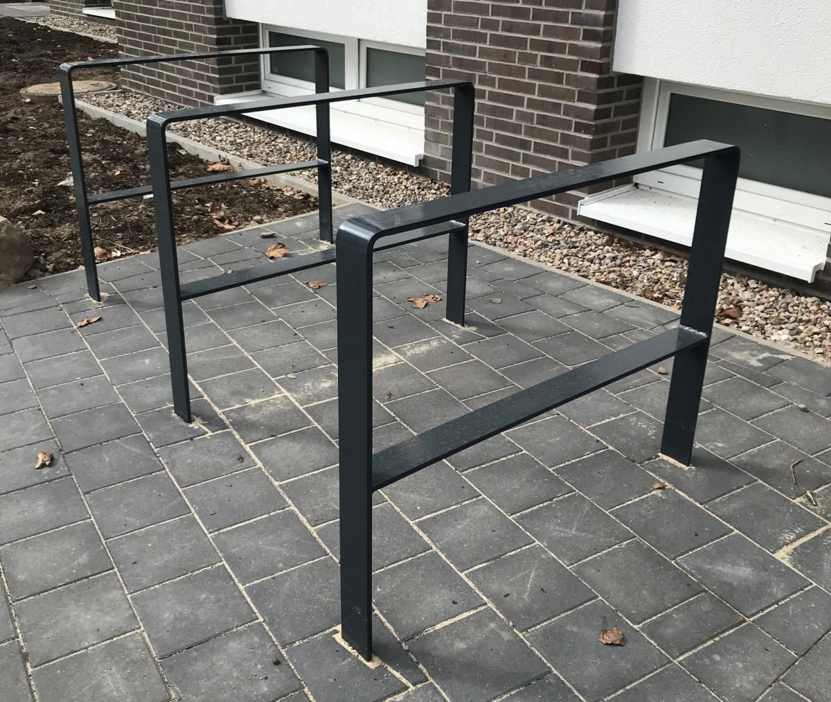 Schwarze Fahrradständer aus Metall auf einem grauen, gepflasterten Gehweg neben einem Backsteingebäude