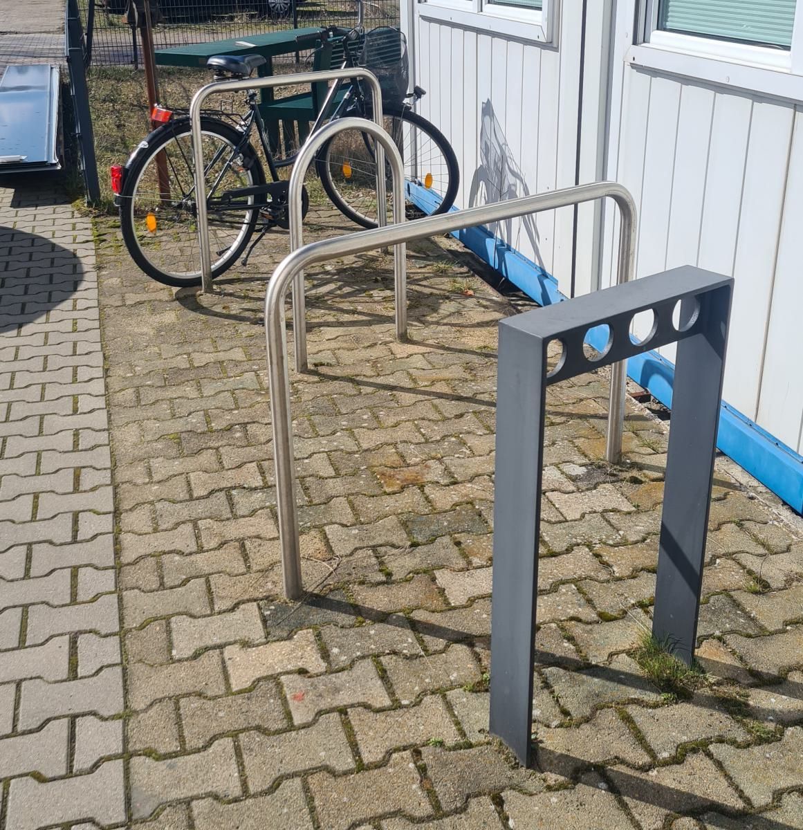 Fahrradständer auf einem gepflasterten Gehweg neben einem weißen Gebäude, im Hintergrund ein geparktes Fahrrad.