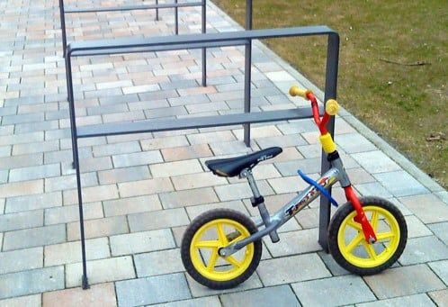 Ein Kinderfahrrad ist an einem Metallständer auf einem gepflasterten Gehweg angeschlossen.