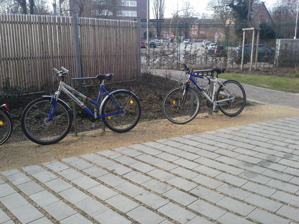 Zwei Fahrräder sind neben einem Zaun auf einem gepflasterten Gehweg in der Nähe einer Rasenfläche abgestellt.