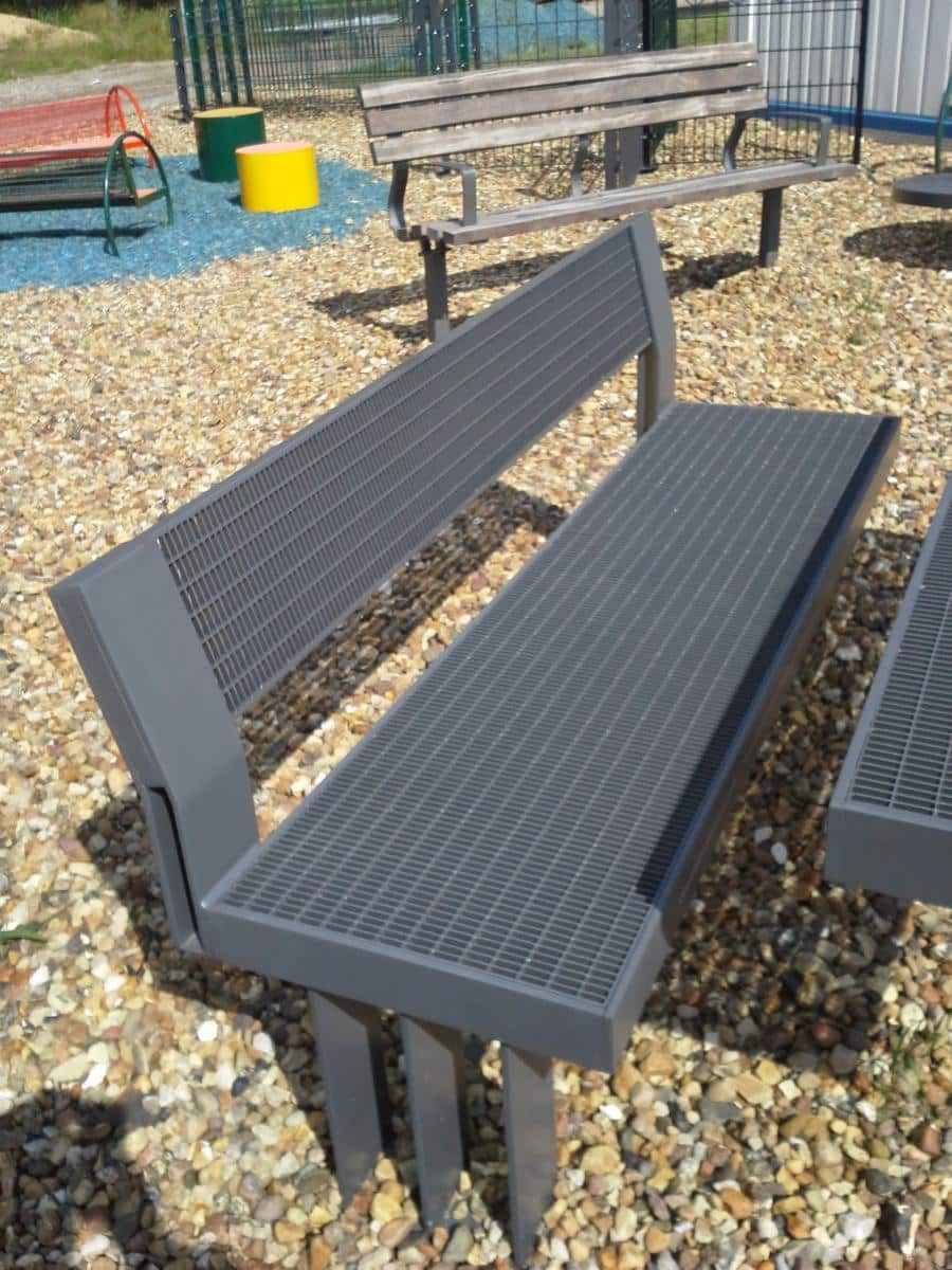 Dunkelgraue Bank auf Kies auf einem Spielplatz, im Hintergrund ein Picknicktisch und Spielgeräte.