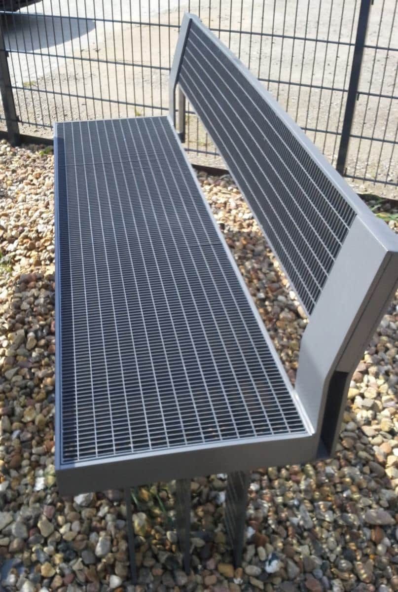 Schwarze Metallbank mit perforierten Sitzflächen auf Kies neben einem Zaun