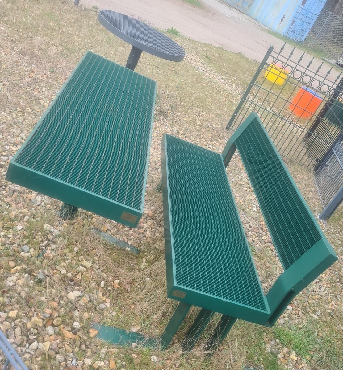 Grüne Picknicktische und Bänke aus Metall auf einer Kiesfläche im Freien in der Nähe eines Zauns