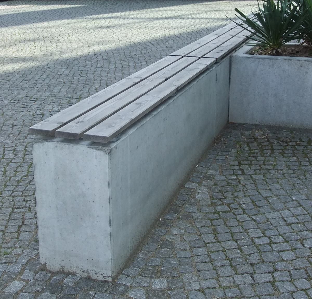 Betonbank mit Holzlatten neben einem gepflasterten Innenhof und Pflanzkübel