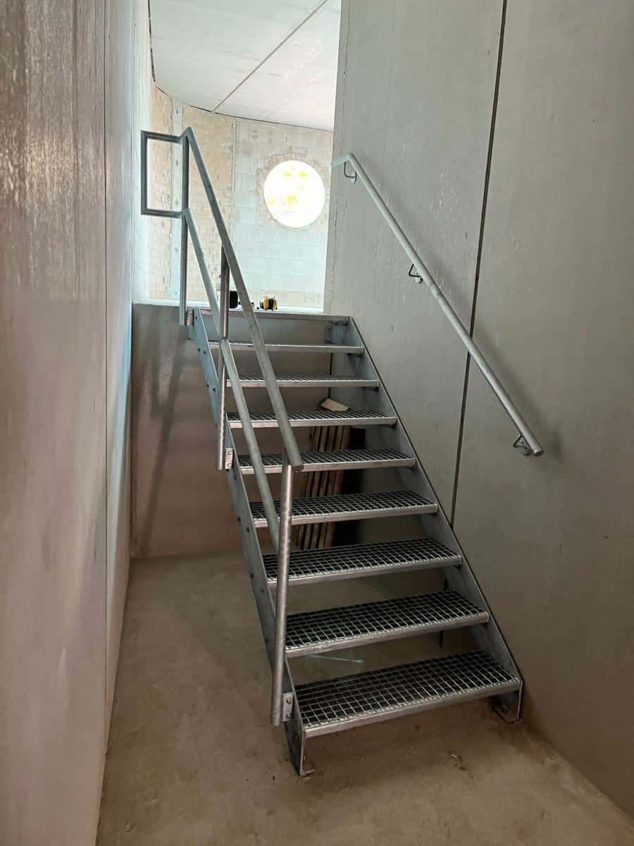 Eine Metalltreppe mit Handlauf in einem schmalen Flur führt zu einem runden Fenster.