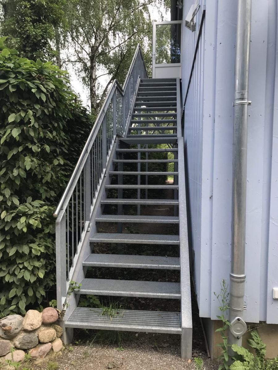 Außentreppe aus Metall neben einem grauen Gebäude, links davon Bäume und Felsen.