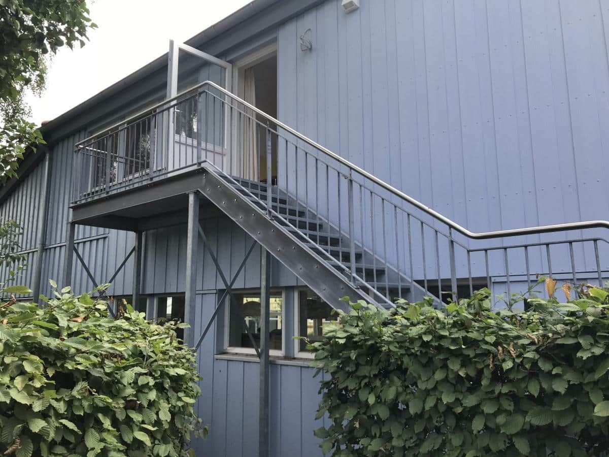 Blaue Gebäudefassade mit Metalltreppe und Balkon, eingerahmt von grünen Sträuchern