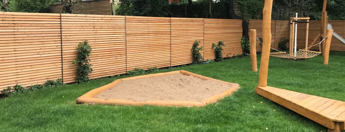 Sandkasten im Garten mit Holzzaun, grünem Rasen und einer erhöhten Terrasse neben Bäumen