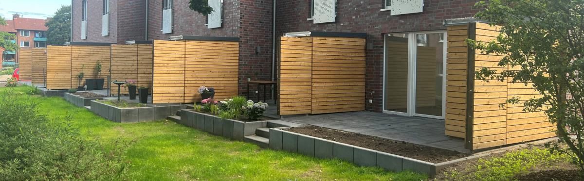 Hinterhofterrasse neben einem Backstein-Reihenhaus, mit Sichtschutzwänden aus Holz und Rasenfläche.