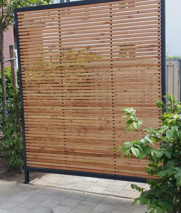 Sichtschutzwand aus Holzlatten, eingefasst in dunkles Metall, neben Pflasterung und Grünflächen.