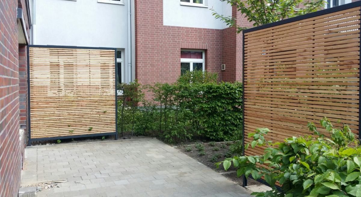 Ein schmaler, gepflasterter Innenhof zwischen Backsteinmauern und Holzlattenzäunen mit Sträuchern und einem kleinen Tor